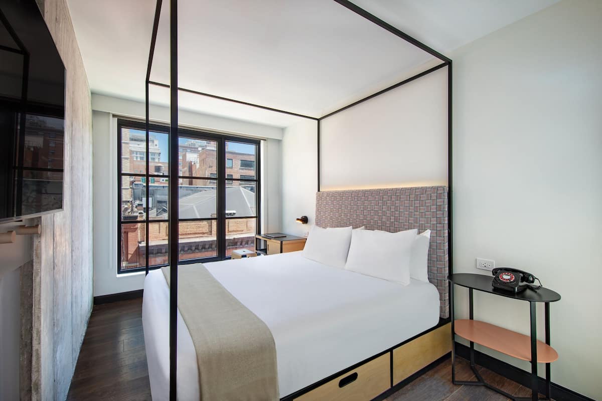 A modern hotel room with a large bed, a small table, and a view of the city through a large window.