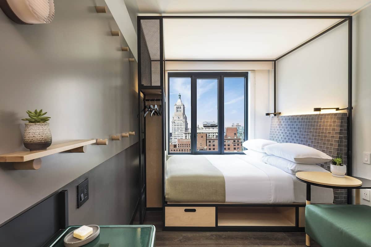A modern hotel room with a large bed, a small table, a chair, a window with a city view, and a shelf with a plant.