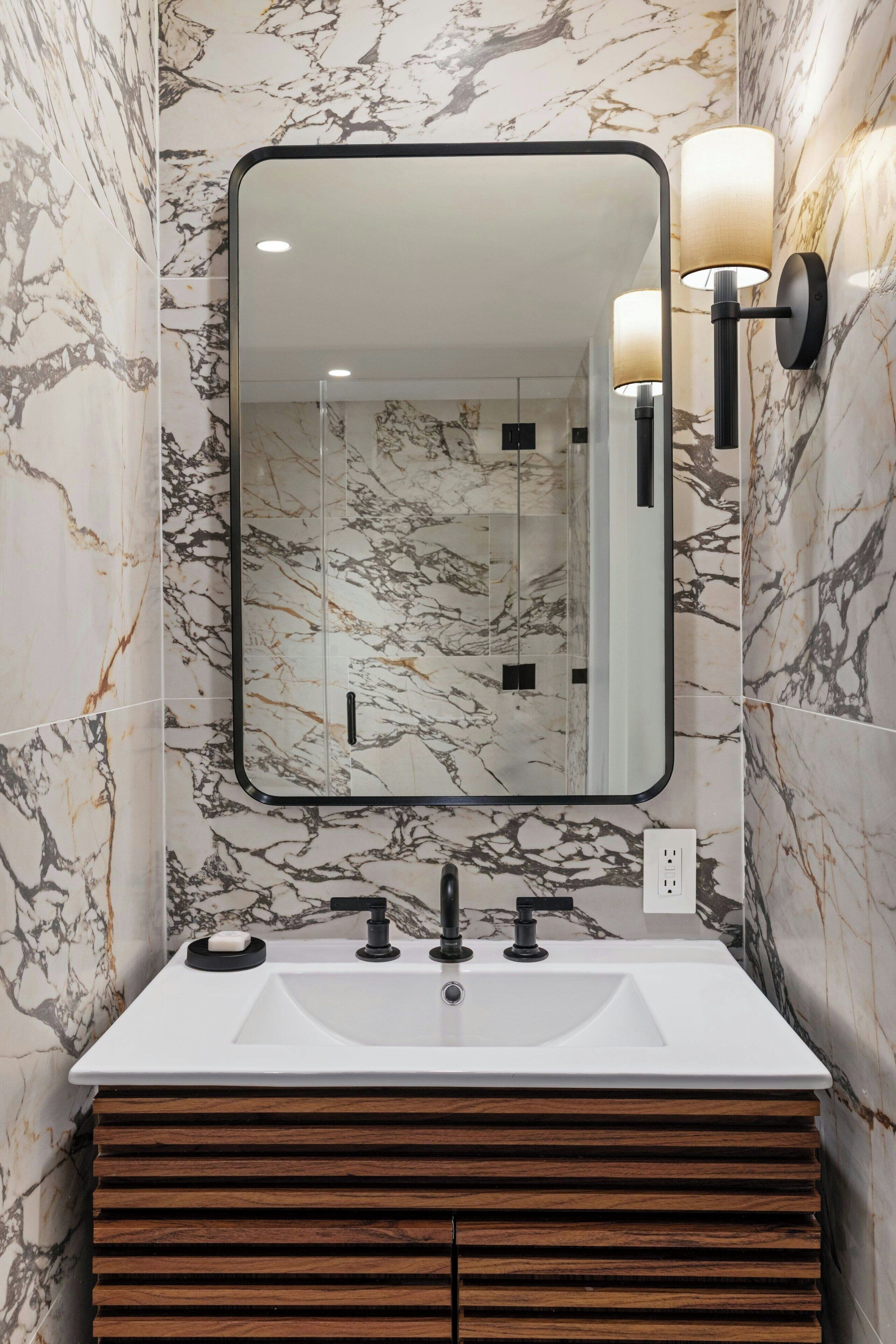 Un baño con pared de mármol, lavamanos blanco, tocador de madera y apliques de pared.