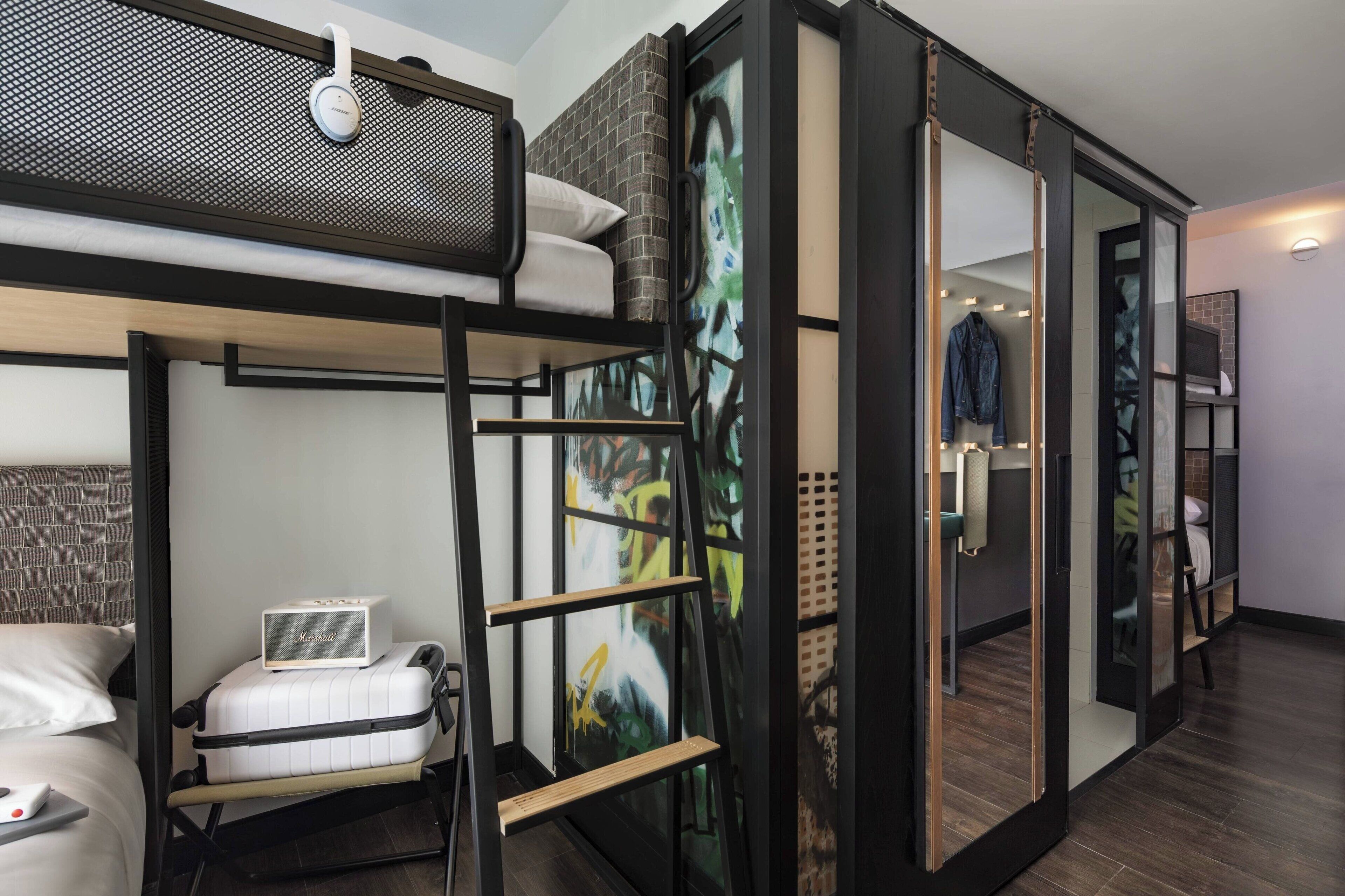 Een moderne hotelkamer met een stapelbed, een Murphy bed, een klein bureau met een laptop en een groot abstract schilderij aan de muur.