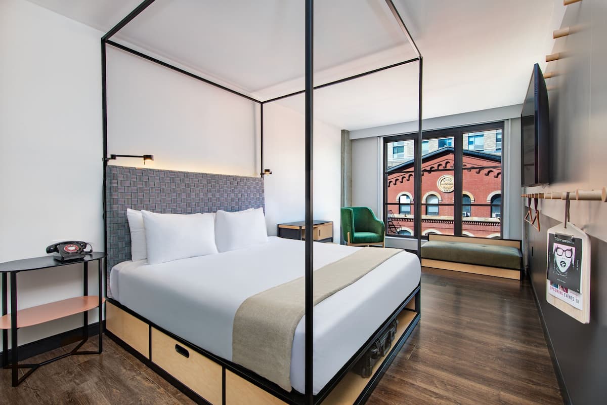 A modern hotel room with a four-poster bed, a small desk with a telephone, a green chair, and a view of a red brick building through the window.