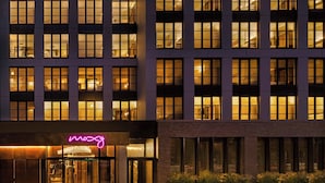 Fachada moderna de hotel con ventanas iluminadas, un letrero de neón y plantas en macetas en el área de entrada.
