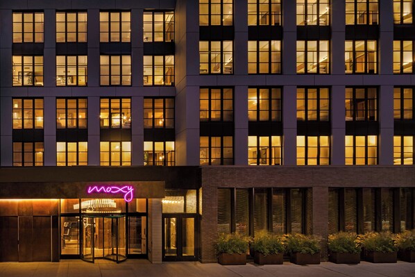 A modern hotel facade with illuminated windows, a neon sign, and potted plants in the entrance area.