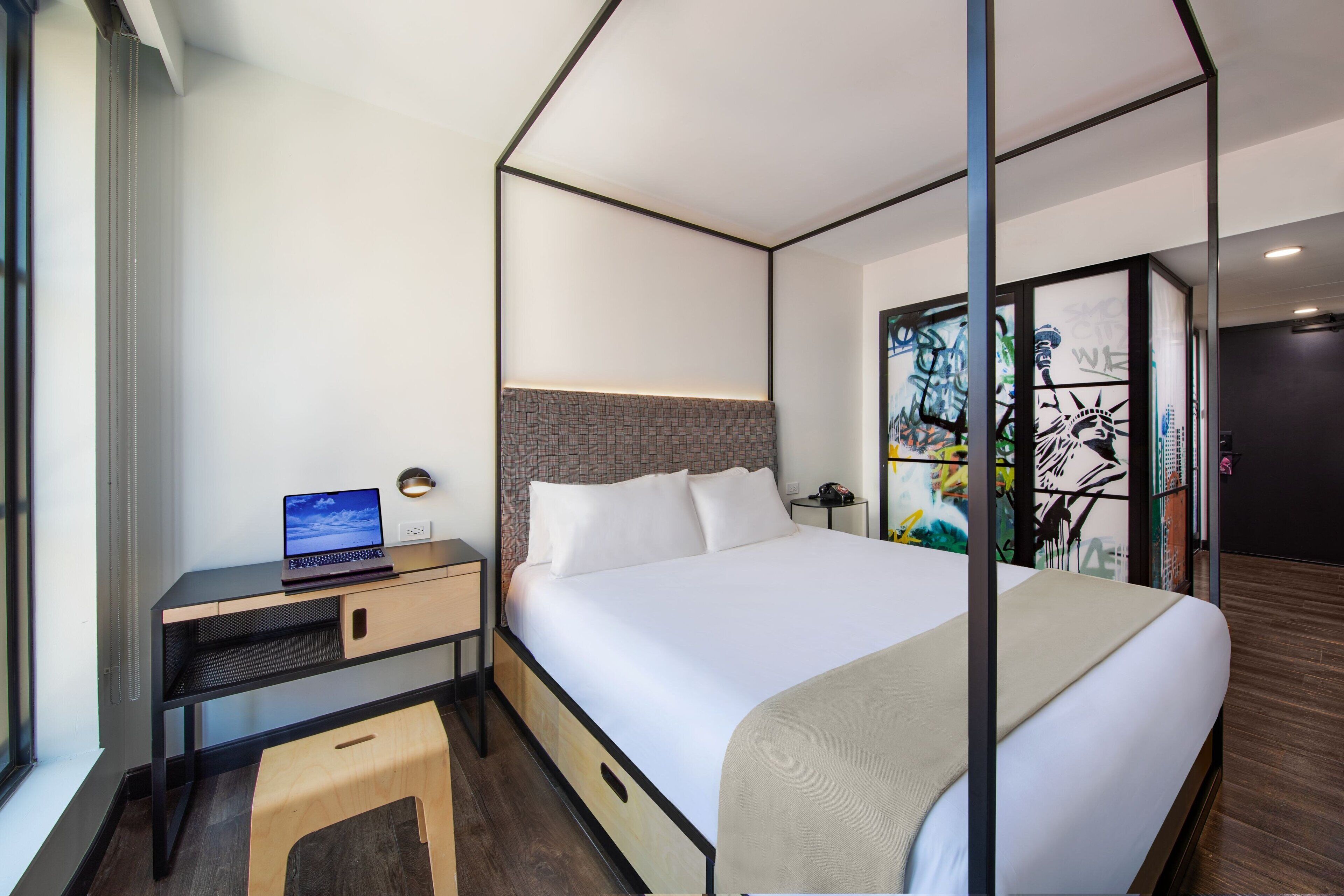 Habitación de hotel moderna con una cama, un escritorio con una computadora portátil y vistas de la ciudad a través de un ventanal.