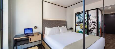 Habitación de hotel moderna con una cama, un escritorio con una computadora portátil y vistas de la ciudad a través de un ventanal.