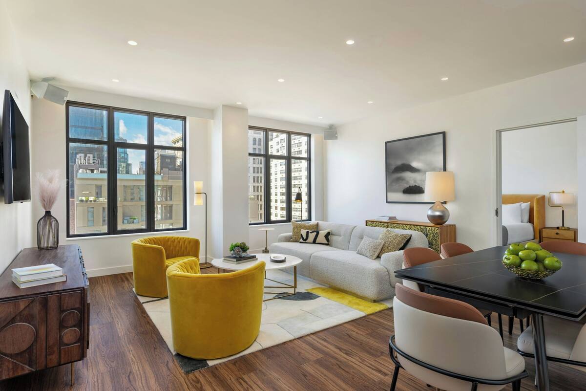 A modern living room with a sofa, yellow chairs, a dining table with apples, and large windows with city views.