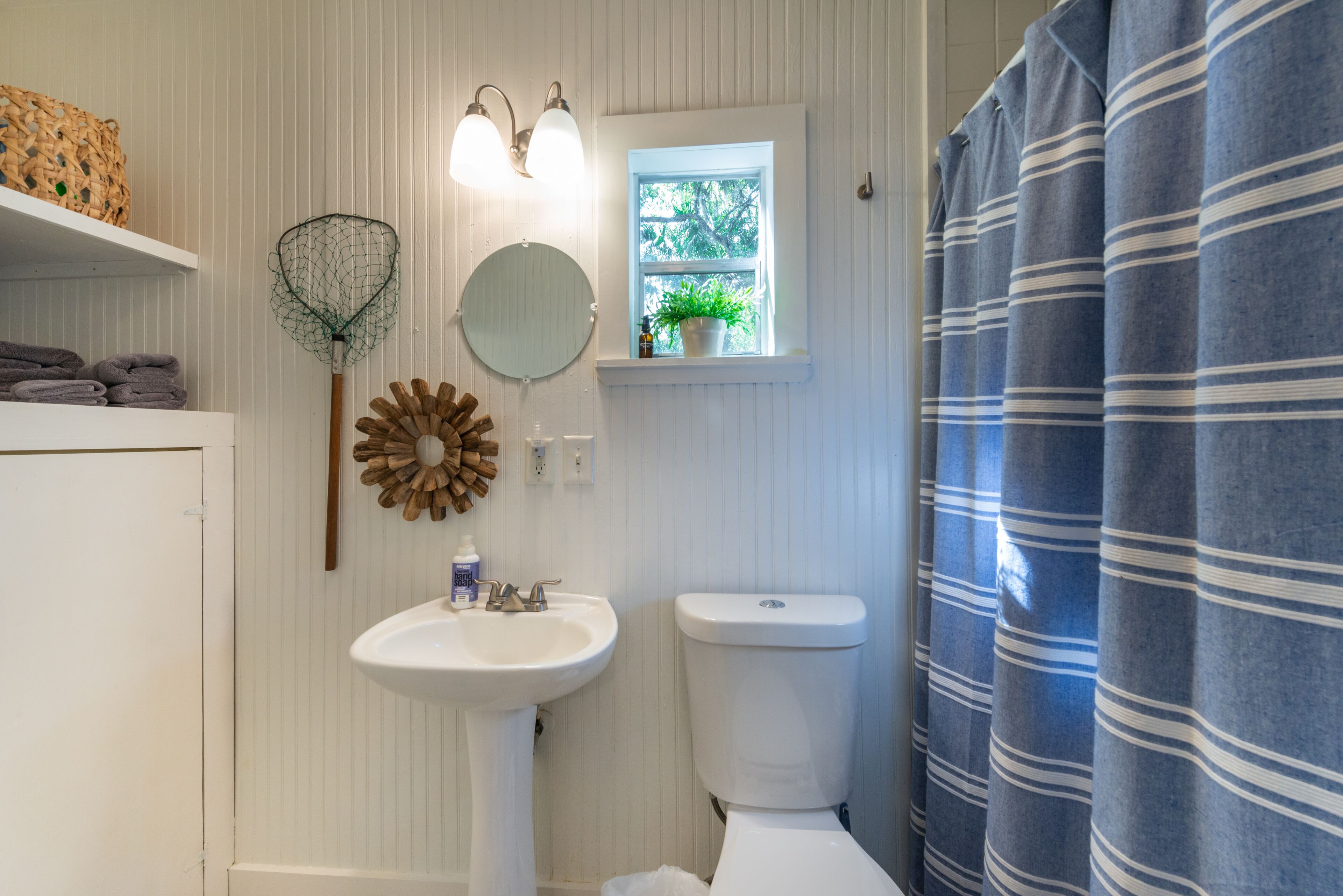 Een badkamer met een wit wastafel, toilet en een raam met een plant. Handdoeken liggen op een plank en er is een douchegordijn.
