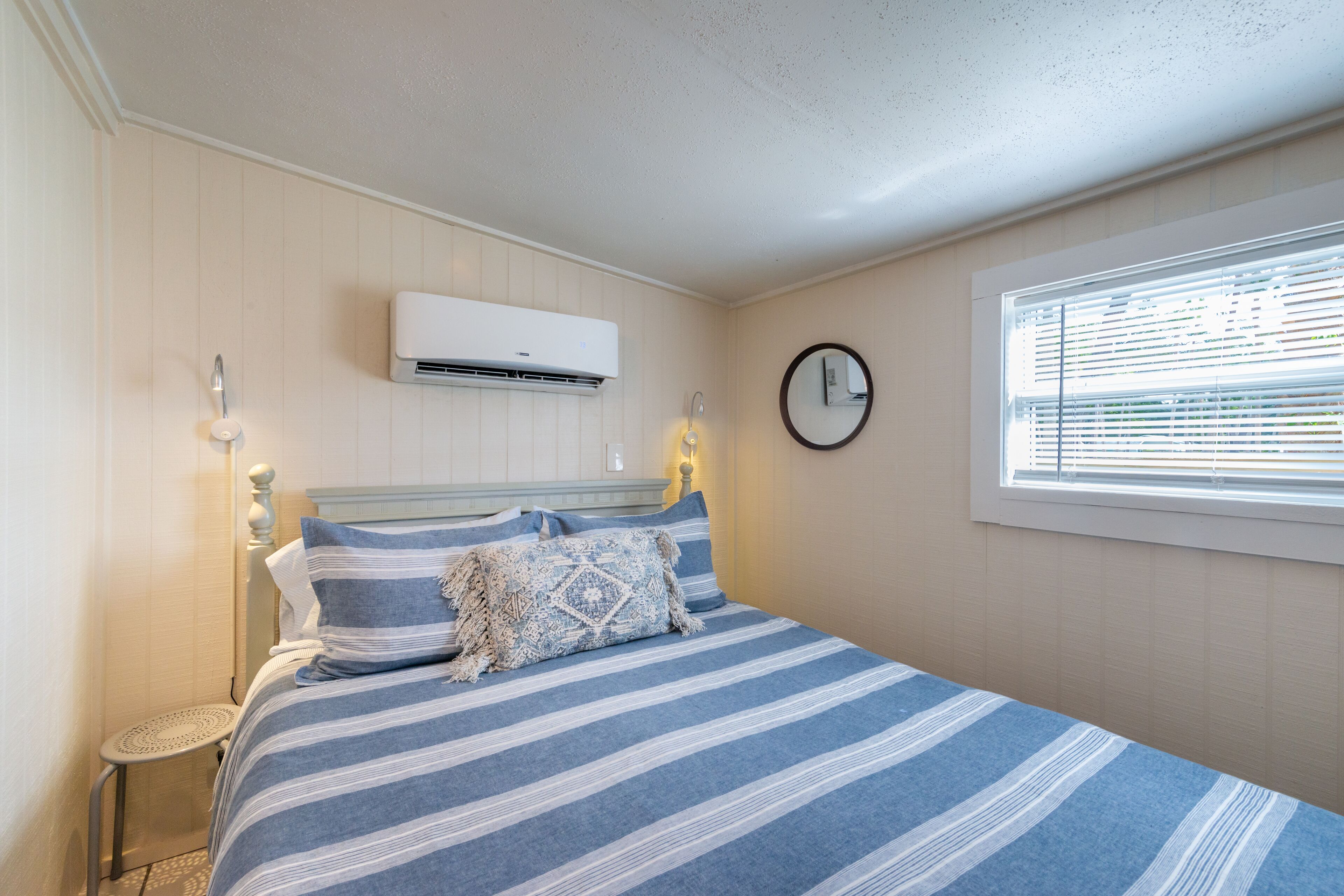 Een slaapkamer met een bed, een nachtkastje, een wandairconditioner, een ronde spiegel en een raam met jaloezieën.