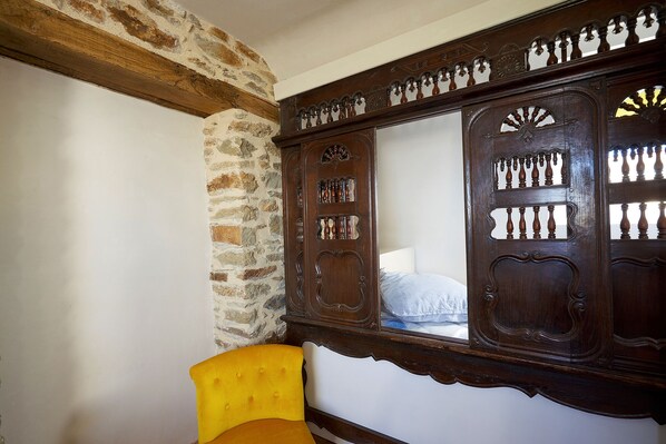 A room with a stone wall, a wooden wardrobe with intricate carvings, and a yellow upholstered chair. - OLD FARM SEA VIEW (Roscoff)