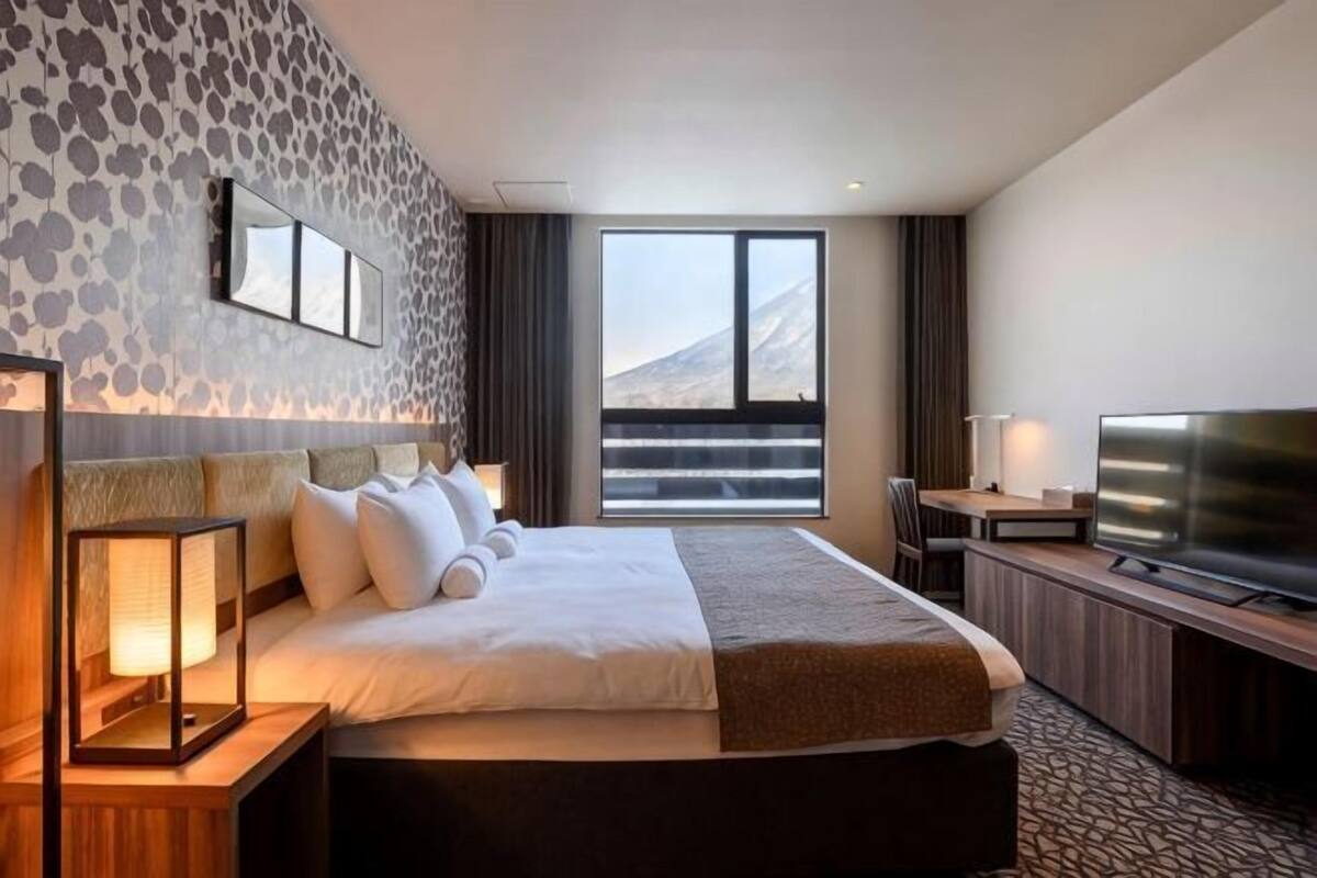 A hotel room with a large bed, a television, a desk with a lamp, and a view of a mountainous landscape.