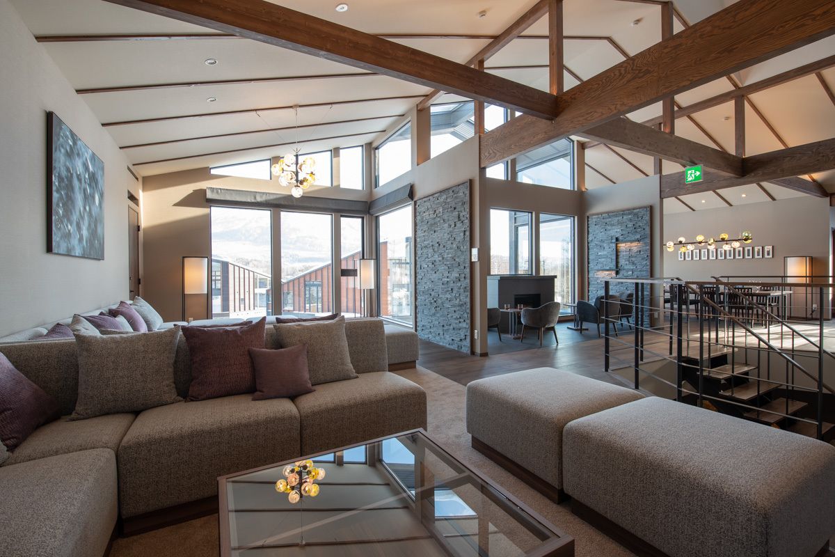 A spacious living area with a high ceiling, exposed beams, a large sofa set, a coffee table, and a view of the city through large windows.