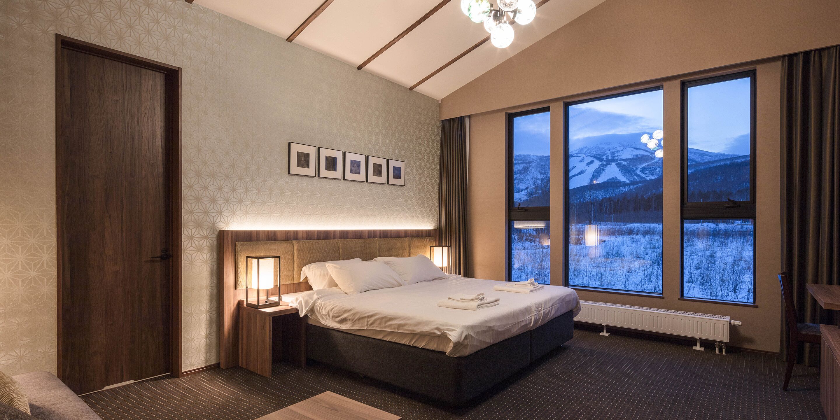 A modern hotel room with a large bed, a nightstand with a lamp, a view of snowy mountains through large windows, and a ceiling with a geometric pattern.