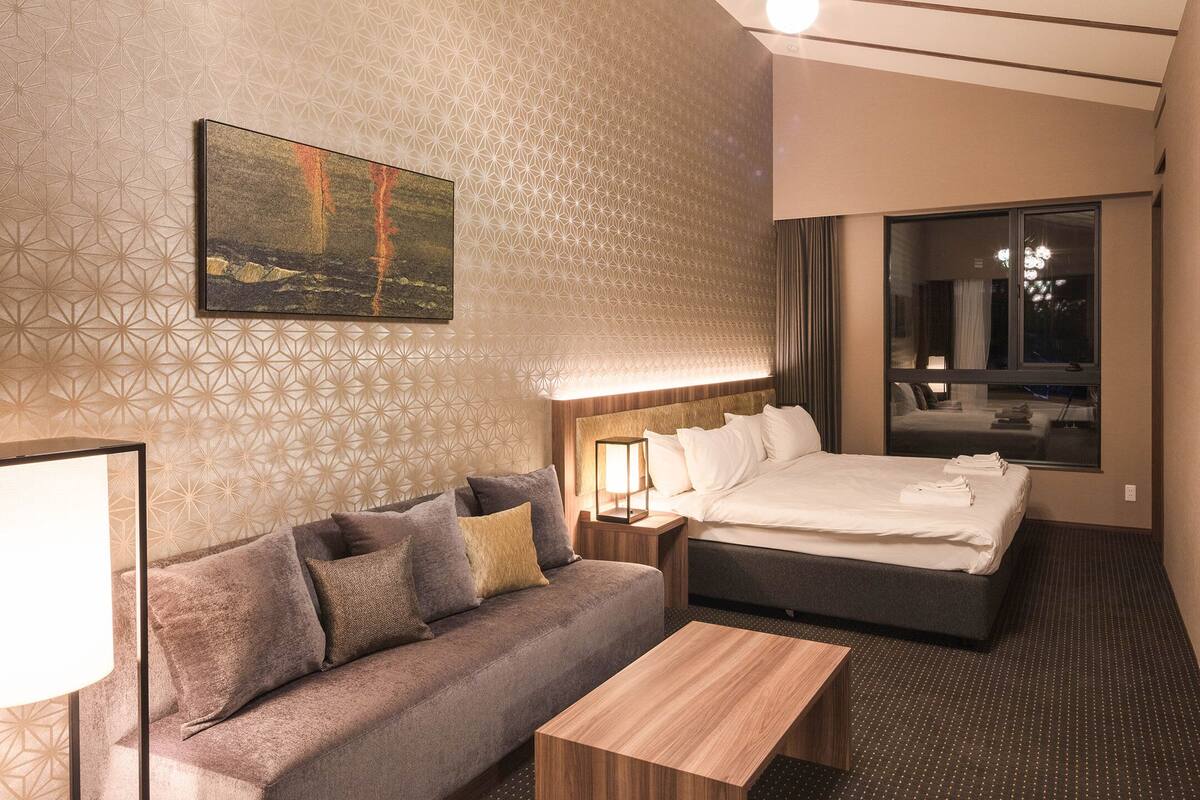 A hotel room with a bed, a sofa, a nightstand, a lamp, and a window.