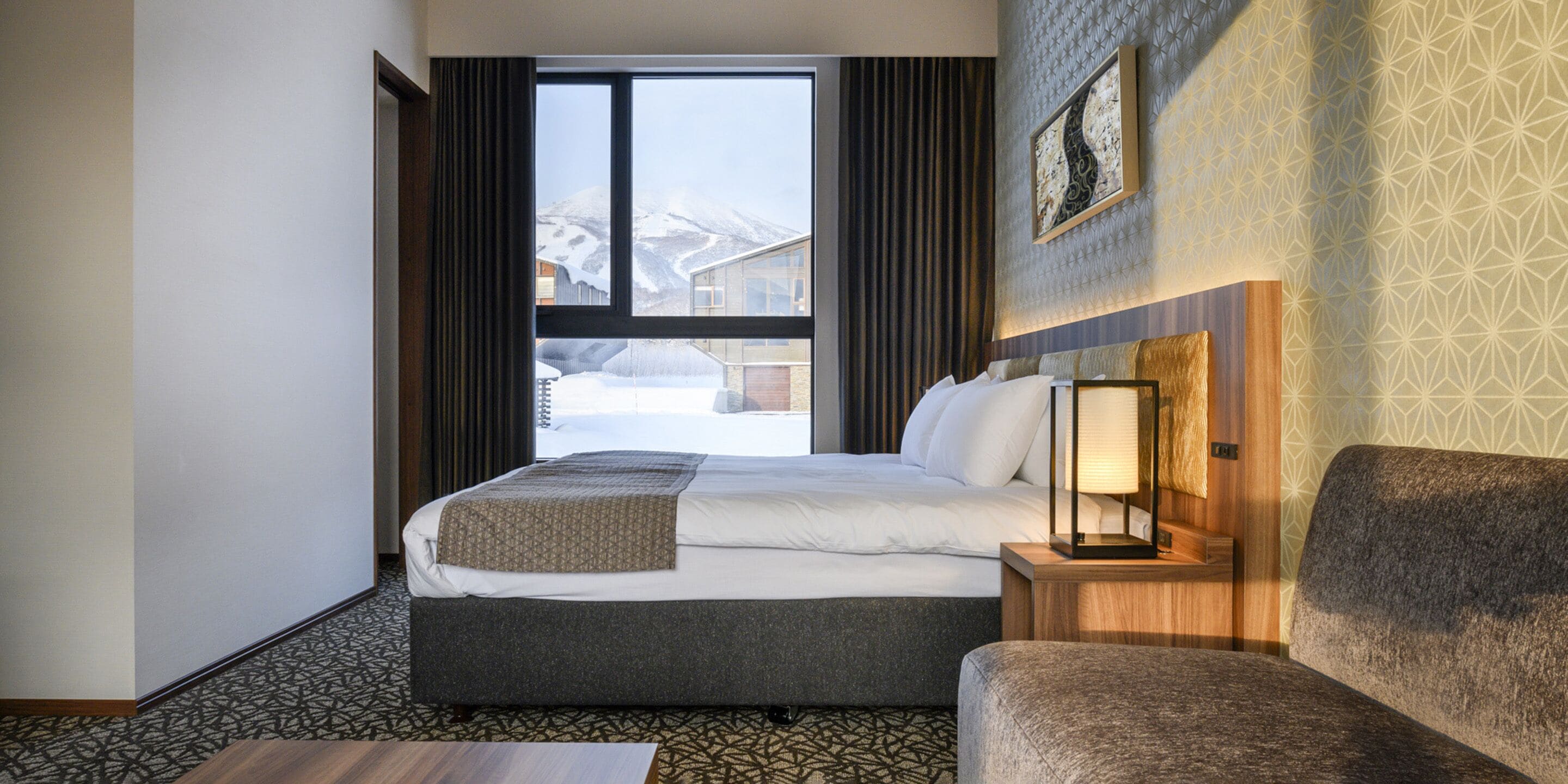 Una habitación de hotel con una cama, una mesita de noche con lámpara, una silla y una ventana con cortinas.