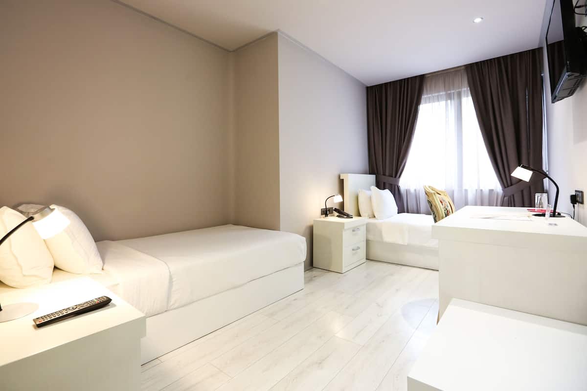 A modern hotel room with a white bed, a desk with a lamp, a chair, and a TV.