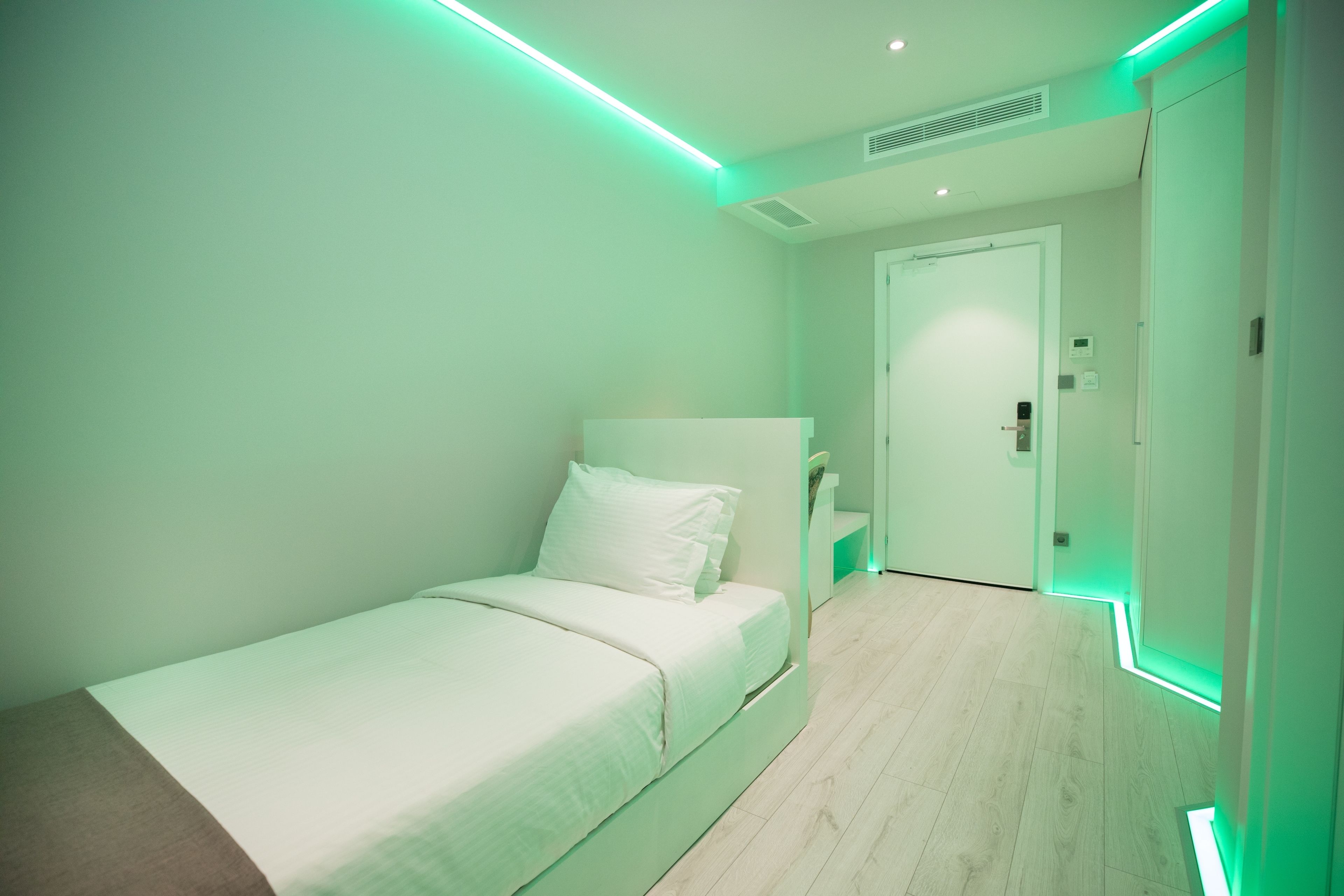 A single-bed room with a wooden floor, a white bed, and green LED lighting along the ceiling.