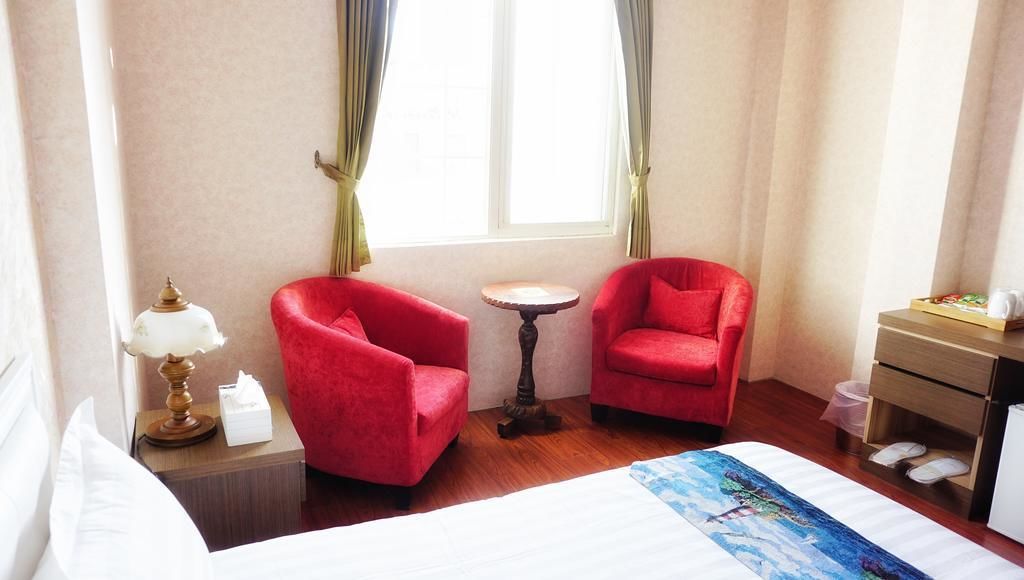 Une chambre d’hôtel avec un lit, deux fauteuils rouges, une petite table ronde, une lampe, une table de chevet, une commode et une fenêtre avec des rideaux.