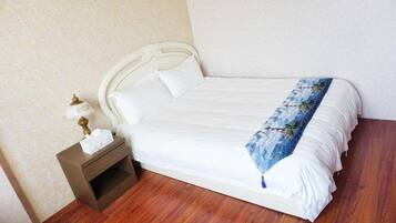 A neatly made bed with white linens and a blue floral throw, a bedside table with a lamp and a tissue box, and a wooden nightstand.