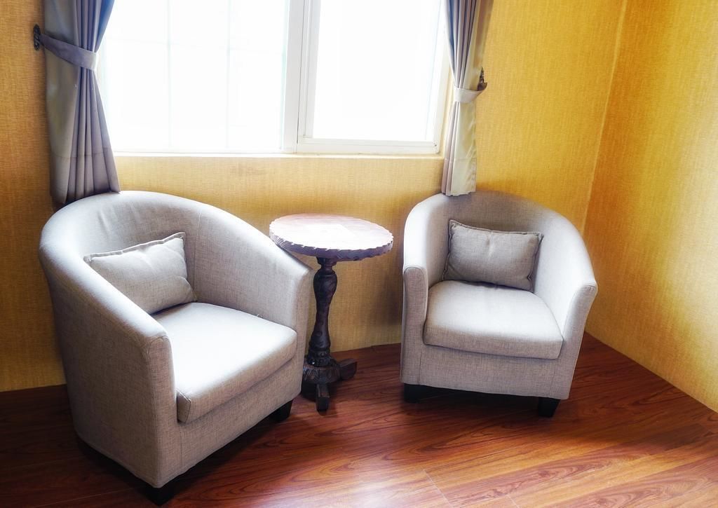 Two armchairs and a small round table in a corner of a room with wooden flooring and a window with curtains.