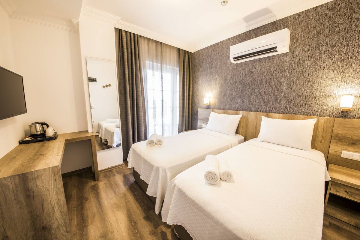 A hotel room with two beds, a wooden desk, a flat-screen TV, an air conditioning unit, and a window with curtains.