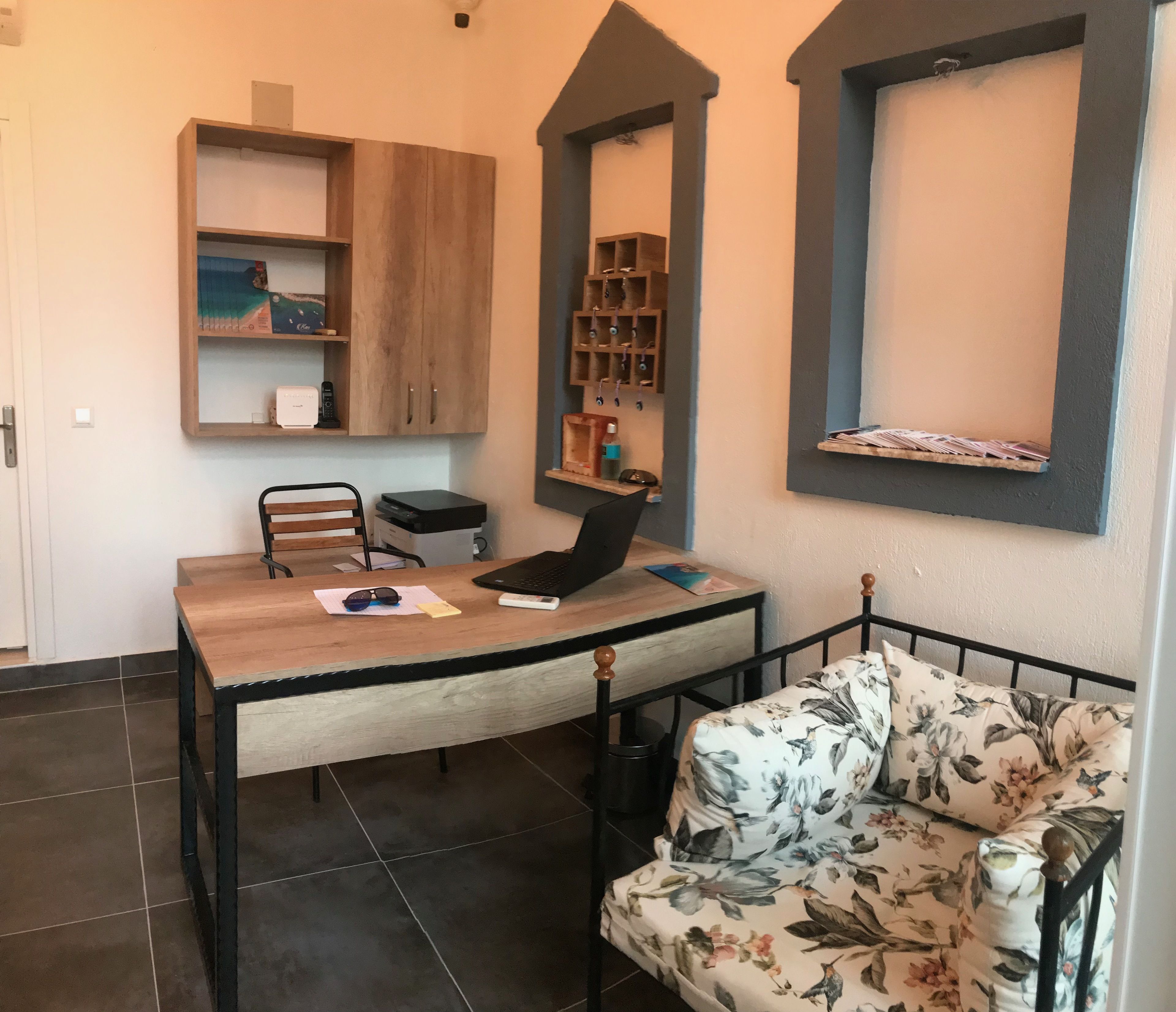 A small, cozy room with a wooden desk, a chair, a laptop, and a floral-patterned sofa. There is a shelf with books and a small cabinet above the desk.
