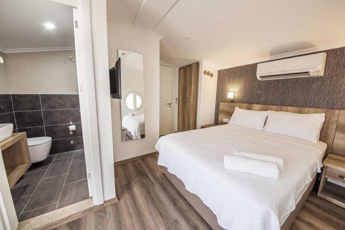 A modern hotel room with a large bed, a wooden headboard, a built-in air conditioning unit, a wooden floor, and a separate bathroom with a toilet and sink.