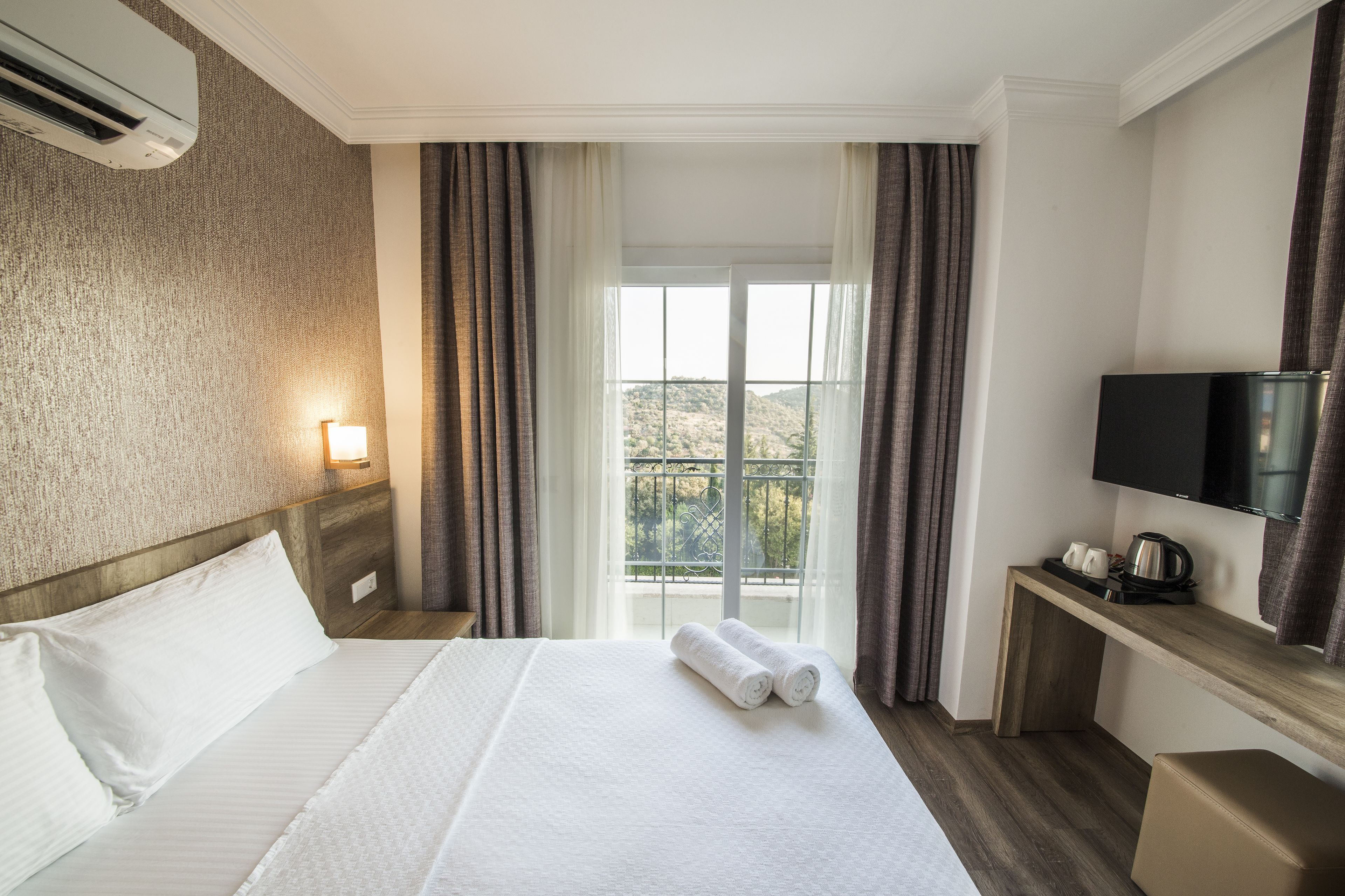 A hotel room with a bed, a TV, a desk with a kettle, and a window with curtains.