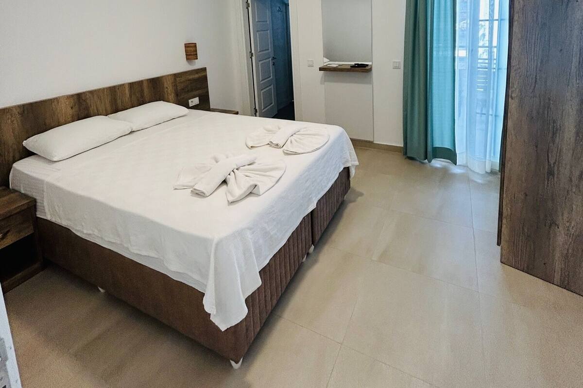 A hotel room with a large bed, a wooden headboard, a nightstand, a mirror, a window with curtains, and a ceiling light.
