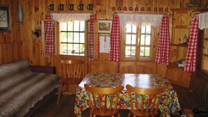 Interior de una cabaña de madera con una mesa de comedor, sillas, un sofá y ventanas con cortinas cuadriculadas.