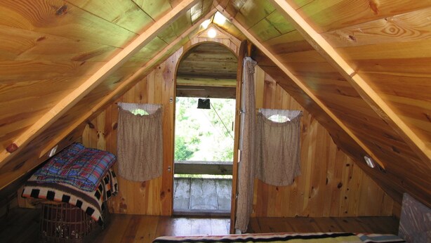 Un dormitorio acogedor en el ático, con techo inclinado, una cama con manta a rayas, una mesita pequeña y cortinas en la ventana.