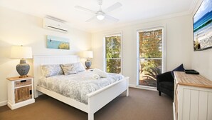 A bedroom with a bed, two bedside tables with lamps, a ceiling fan, and a large window with blinds. - Calming Waters - Pet Friendly - Community Pool (Callala Beach)