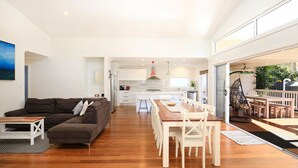 A modern living room with a dark brown sofa, a wooden dining table with white chairs, and a kitchen in the background. - Calming Waters - Pet Friendly - Community Pool (Callala Beach)
