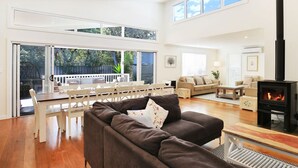 A spacious living room with a large sofa, a dining area with a table and chairs, a fireplace, and large windows providing natural light. - Calming Waters - Pet Friendly - Community Pool (Callala Beach)