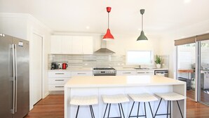 A modern kitchen with white cabinetry, a stainless steel refrigerator, a gas stove, and a large island with bar stools. There are two pendant lights, one red and one green. - Calming Waters - Pet Friendly - Community Pool (Callala Beach)