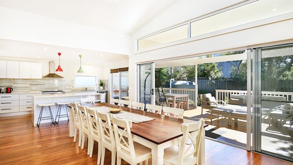 A modern kitchen with white cabinets, a wooden dining table, and a large sliding glass door leading to a deck. - Calming Waters - Pet Friendly - Community Pool (Callala Beach)