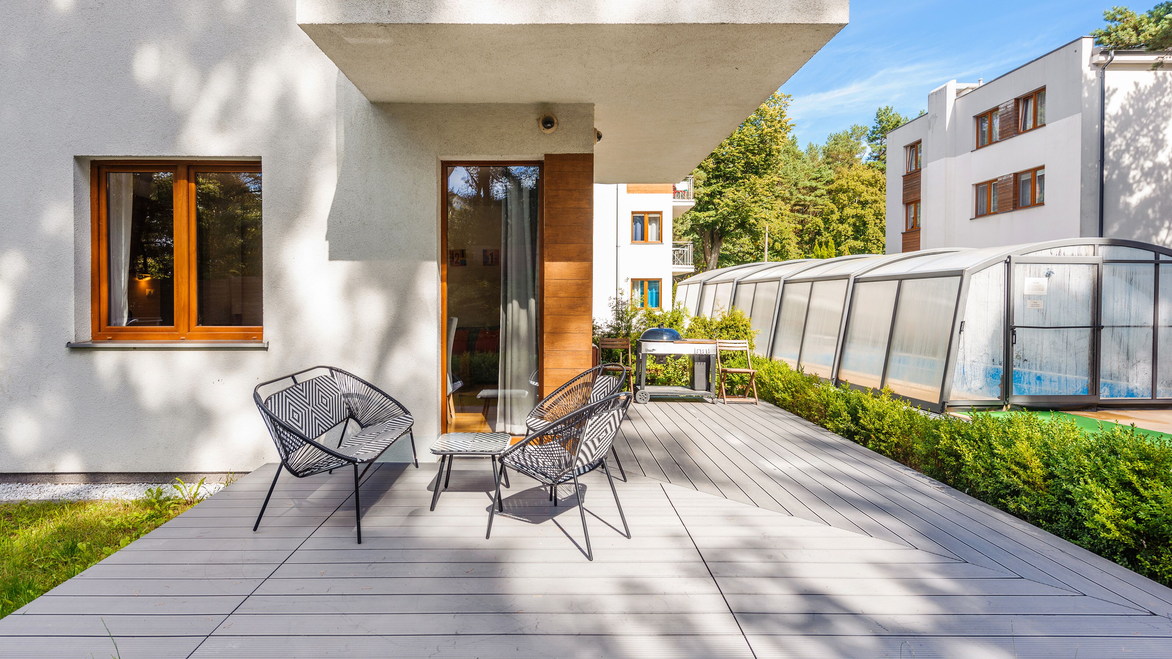 Un patio extérieur moderne avec deux chaises en métal et une petite table, une terrasse en bois et une structure de serre.