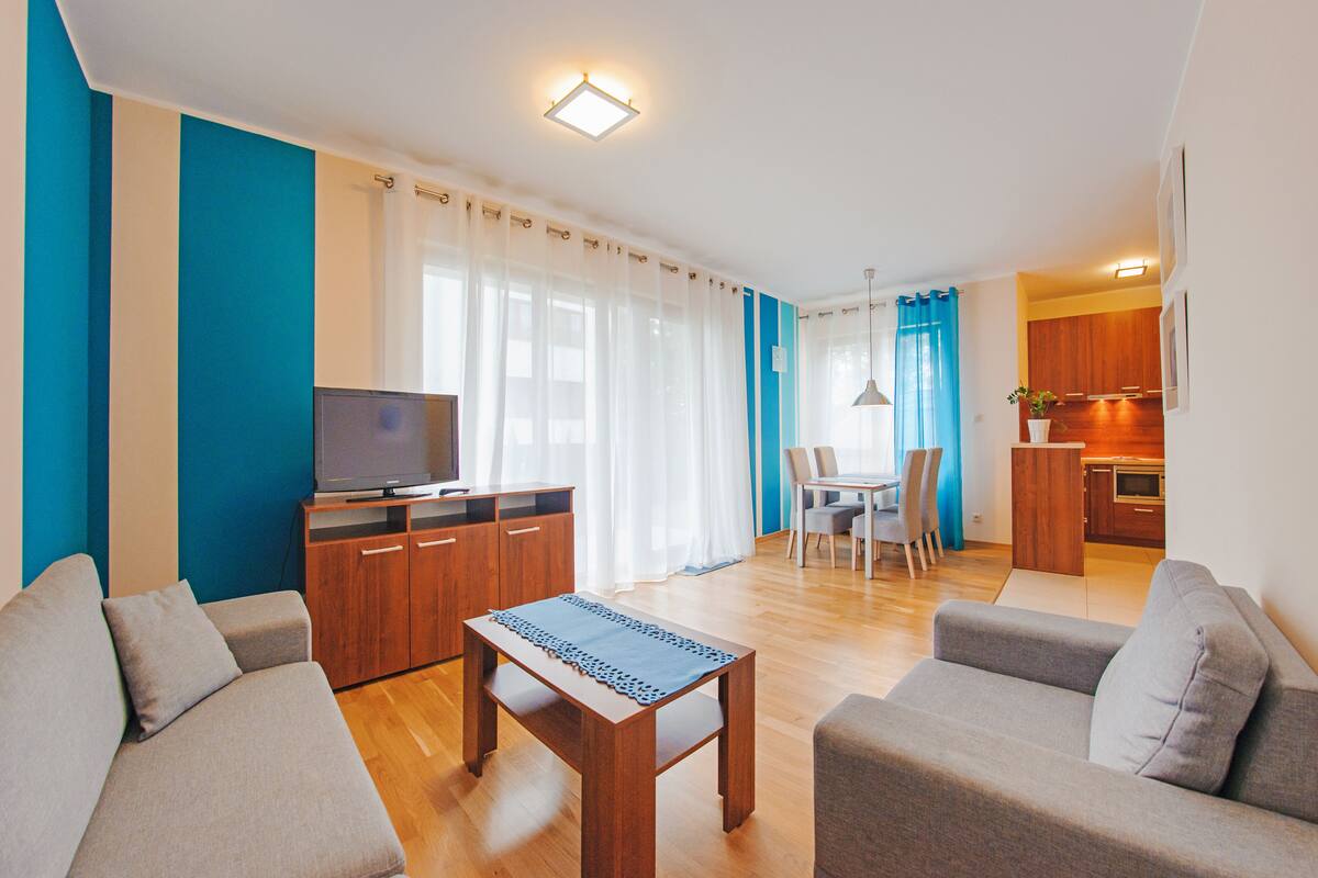 A modern living room with a sofa, a coffee table, a TV on a wooden cabinet, and a dining area with chairs and a table.