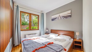 A bedroom with a wooden bed, a window with curtains, a radiator, a bedside table with a lamp, and a wall-mounted picture. - Apartamenty Sun & Snow Cztery Pory Roku (Wladyslawowo)