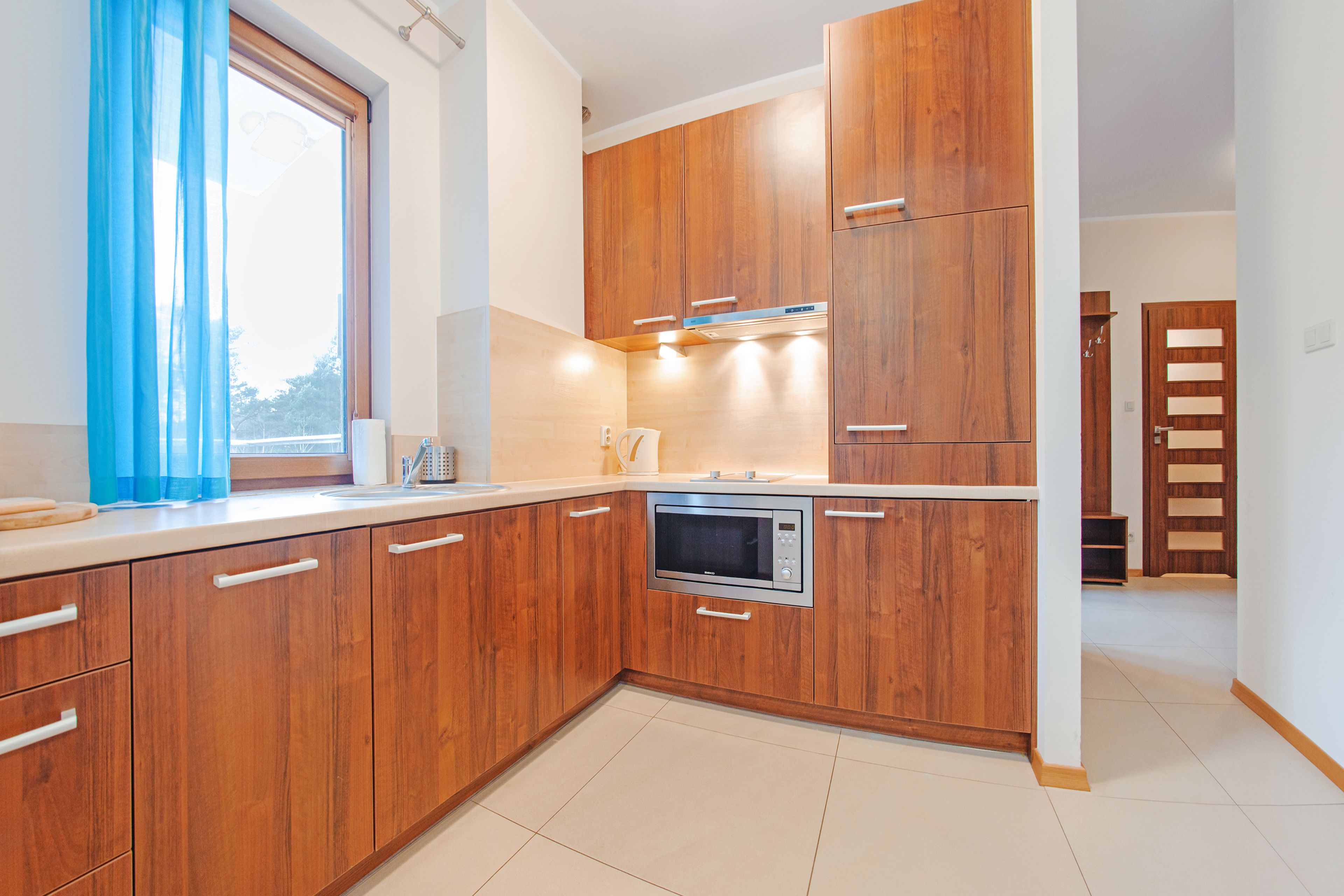 A modern kitchen with wooden cabinets, a built-in microwave, and a window with blue curtains.