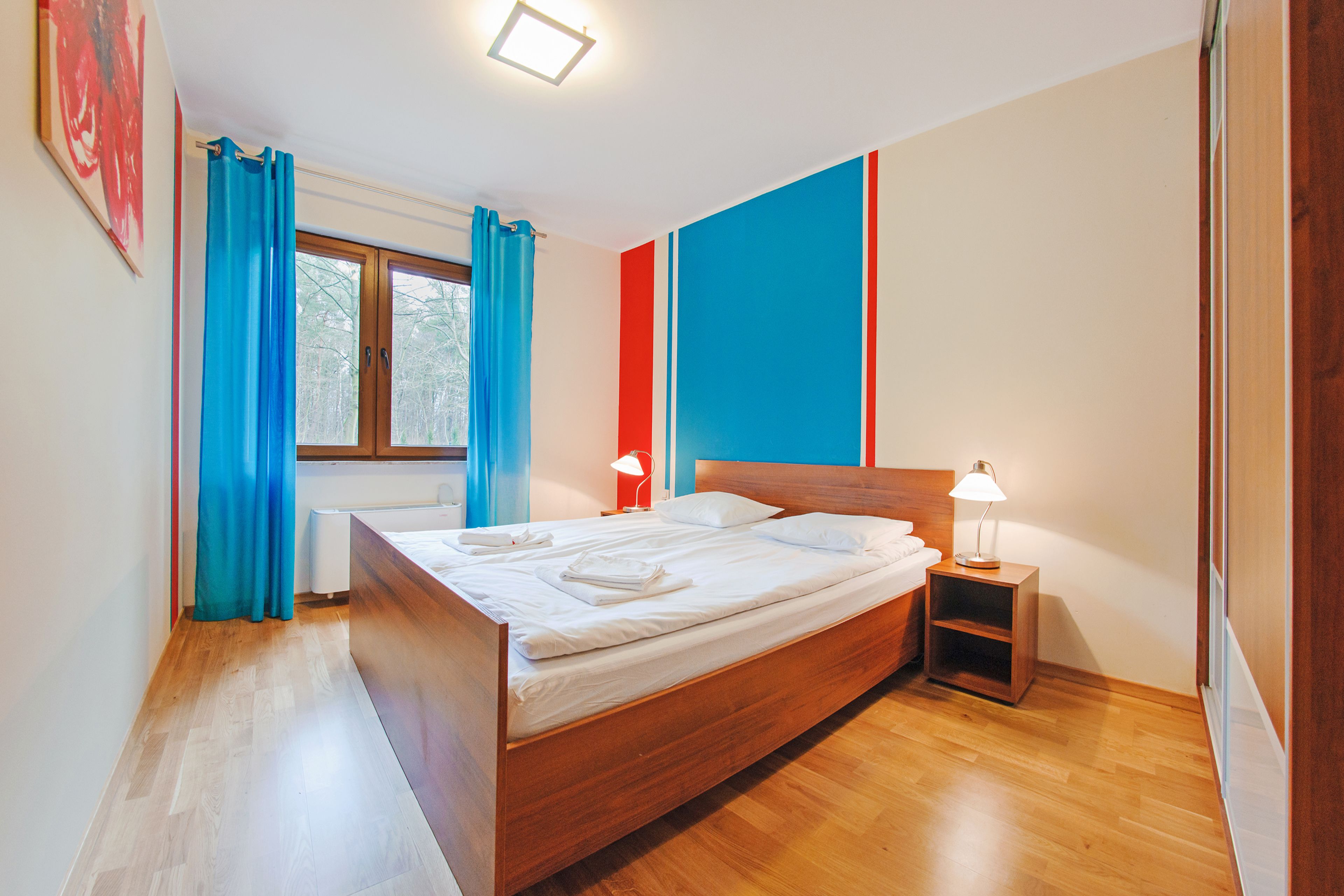 A bedroom with a wooden bed, a nightstand with a lamp, and a window with curtains.