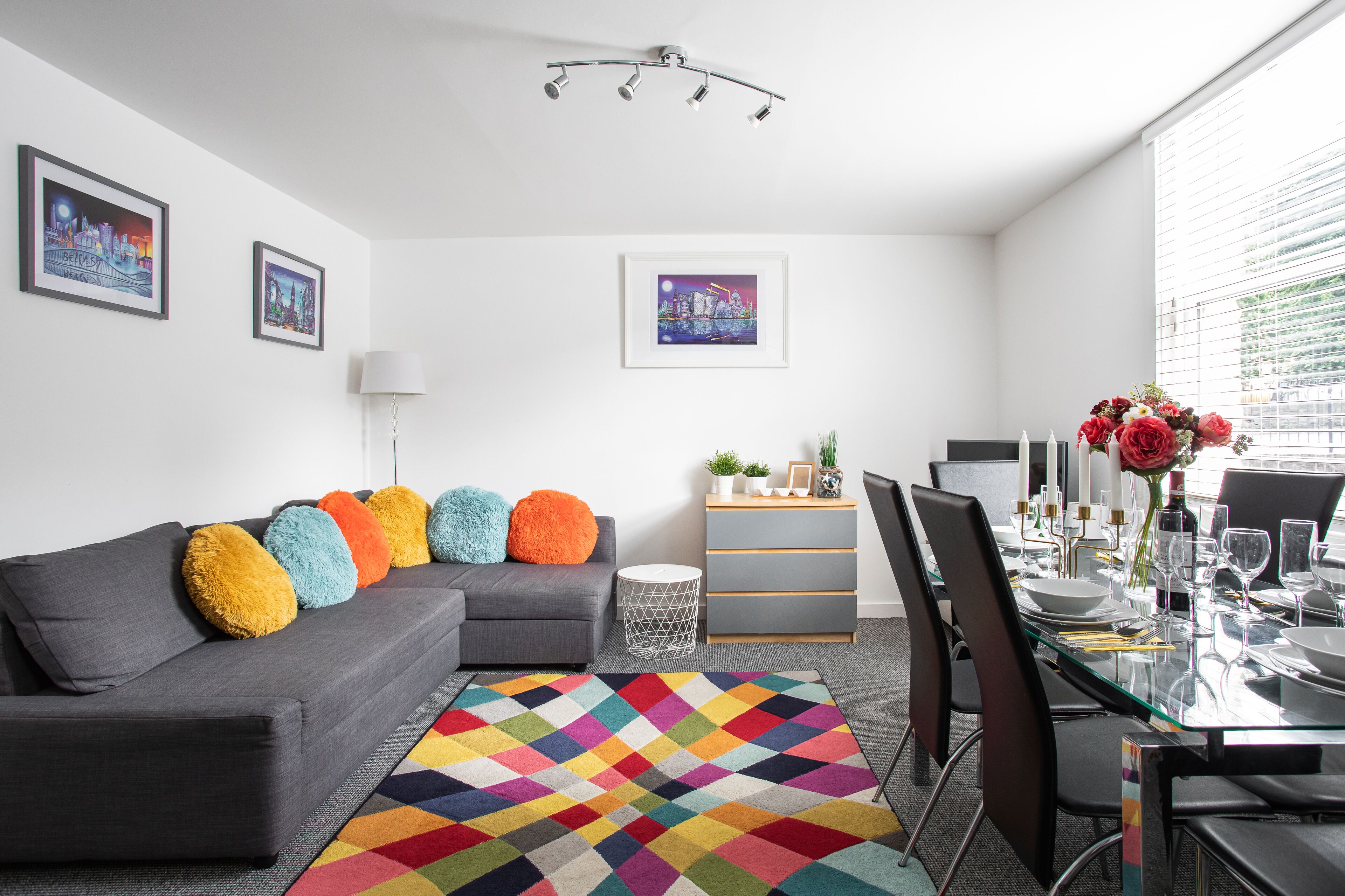 A modern living room with a gray sofa, colorful pillows, a glass dining table set for six, and a cityscape painting on the wall.