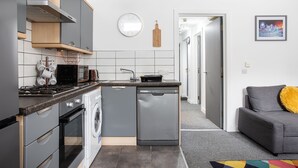Une cuisine compacte équipée d’un lave-vaisselle, d’une cuisinière et d’un four à micro-ondes. Le salon comprend un canapé gris agrémenté d’un coussin jaune.