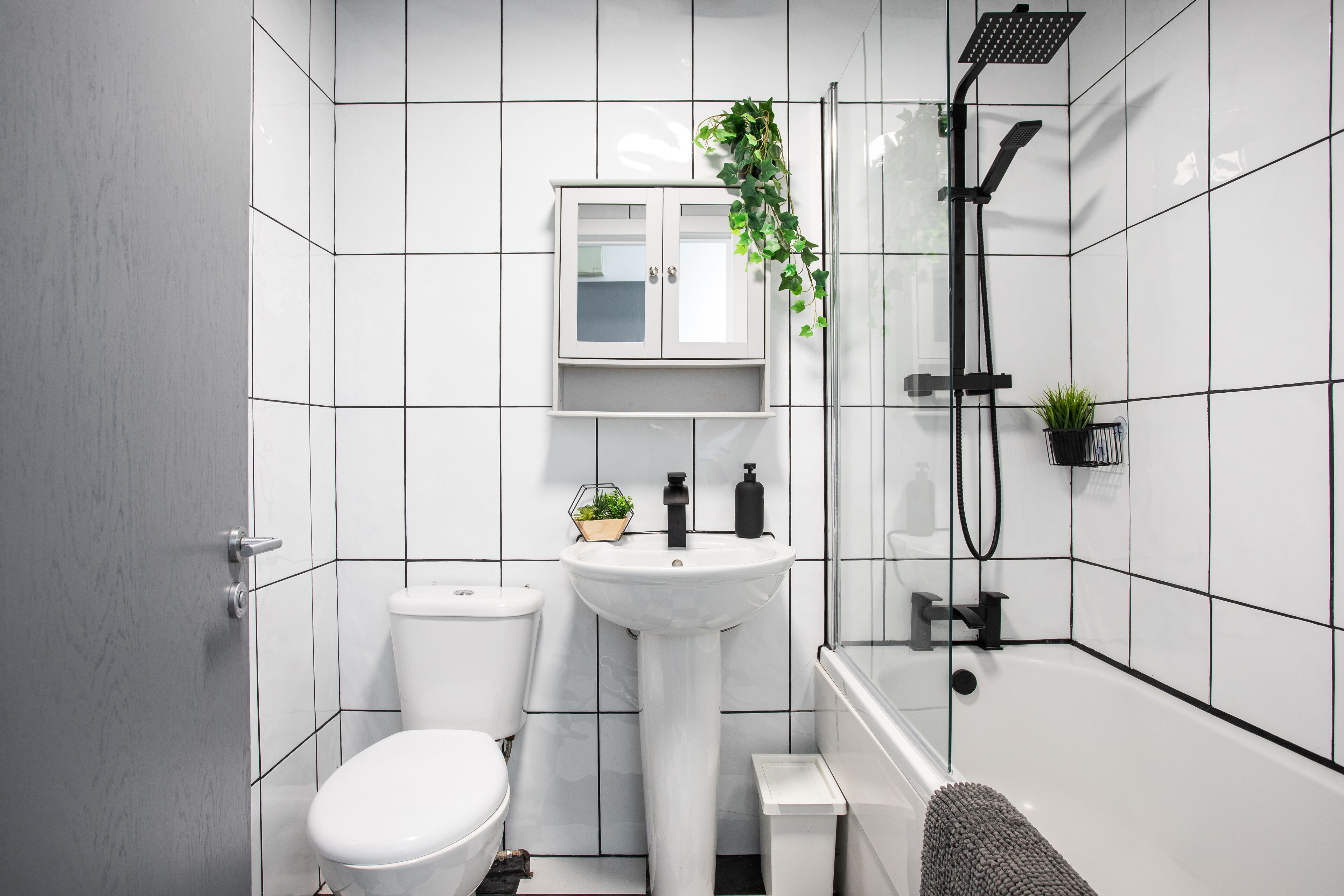Un baño moderno con baldosas blancas, un lavamanos blanco, un inodoro, una ducha y un mueble con una planta.