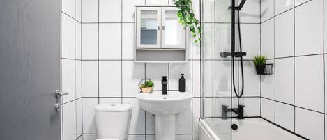 Un baño moderno con baldosas blancas, un lavamanos blanco, un inodoro, una ducha y un mueble con una planta.