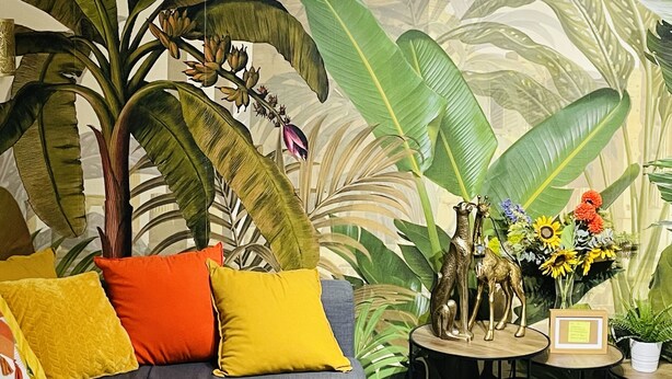 Une pièce décorée d’une grande fresque murale aux motifs tropicaux, avec un canapé gris agrémenté de coussins jaunes et orange, et une petite table sur laquelle sont disposés des objets décoratifs et un tableau encadré.