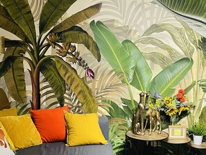 Une pièce décorée d’une grande fresque murale aux motifs tropicaux, avec un canapé gris agrémenté de coussins jaunes et orange, et une petite table sur laquelle sont disposés des objets décoratifs et un tableau encadré.