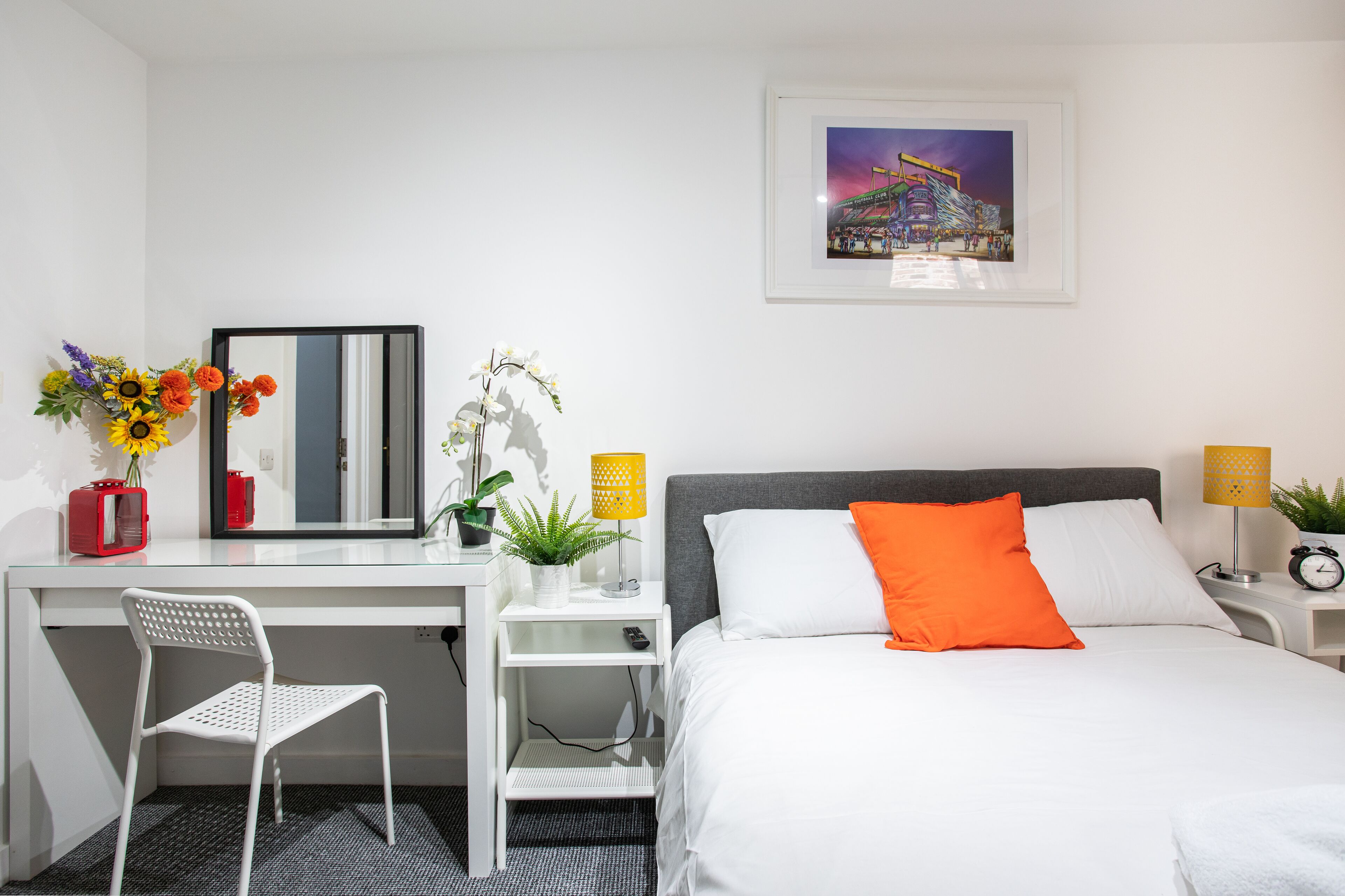 A hotel room with a bed, a desk, a chair, a mirror, a framed picture on the wall, and a small table with a lamp and a clock.