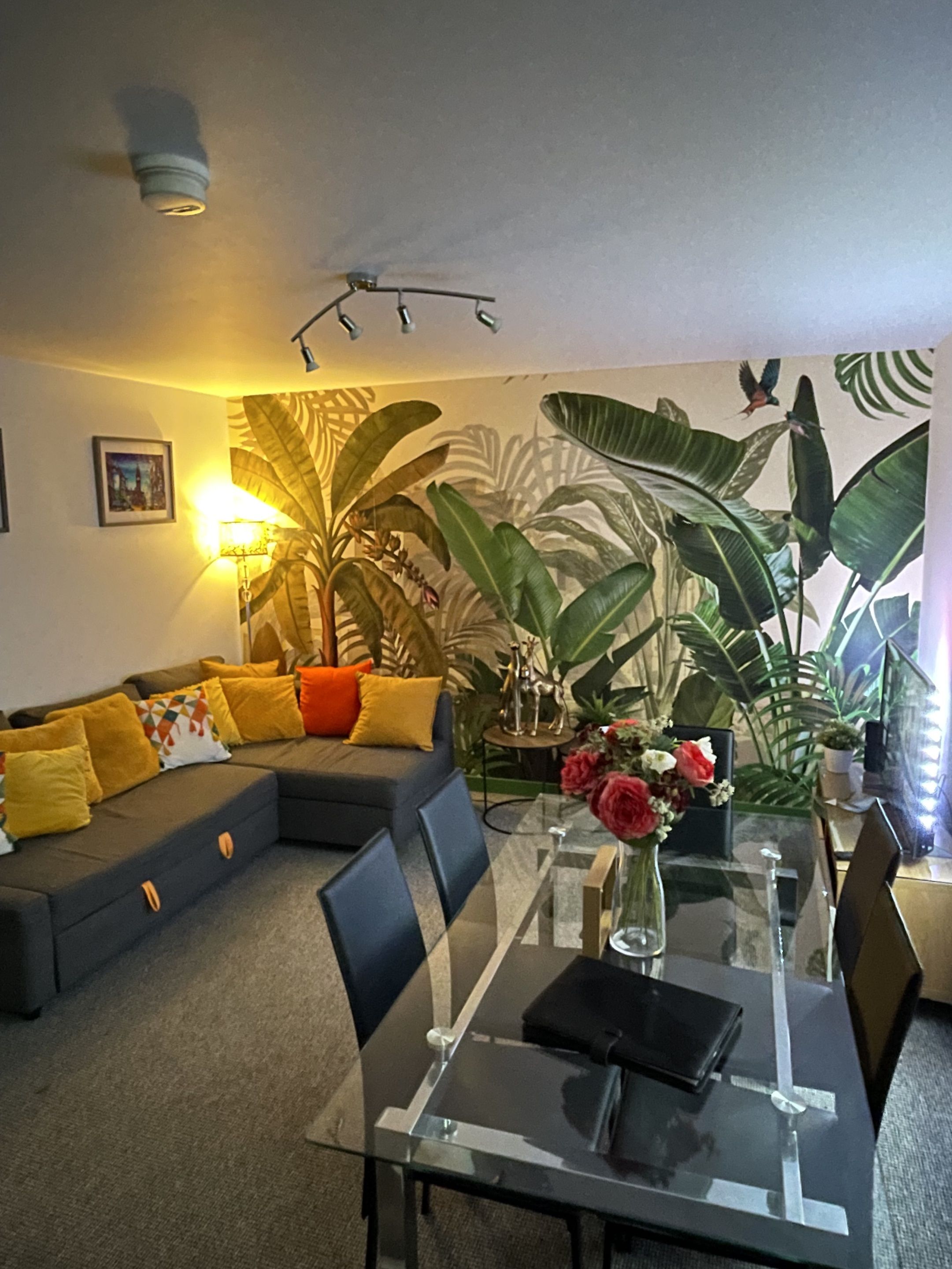 Ein modernes Wohnzimmer mit einem Glastisch, einem Ecksofa mit gelben Kissen, einer Topfpflanze und einer Wandleuchte.
