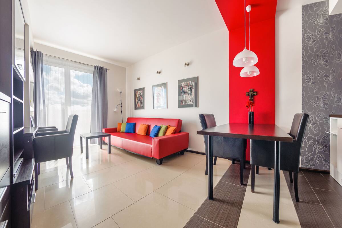 A modern living room with a red sofa, a dining table, and black chairs. There are three framed artworks on the wall and a cross decoration.