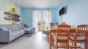 A living room with a sofa, a dining table with chairs, a TV mounted on the wall, and a view of the outdoors through a glass door. - Apartamenty Sun & Snow Lisi Jar (Wladyslawowo)